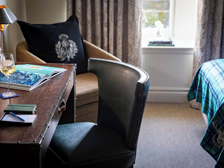 A hotel room with a wooden desk, a leather chair, a bed with a plaid blanket, a window with curtains, and a lamp.