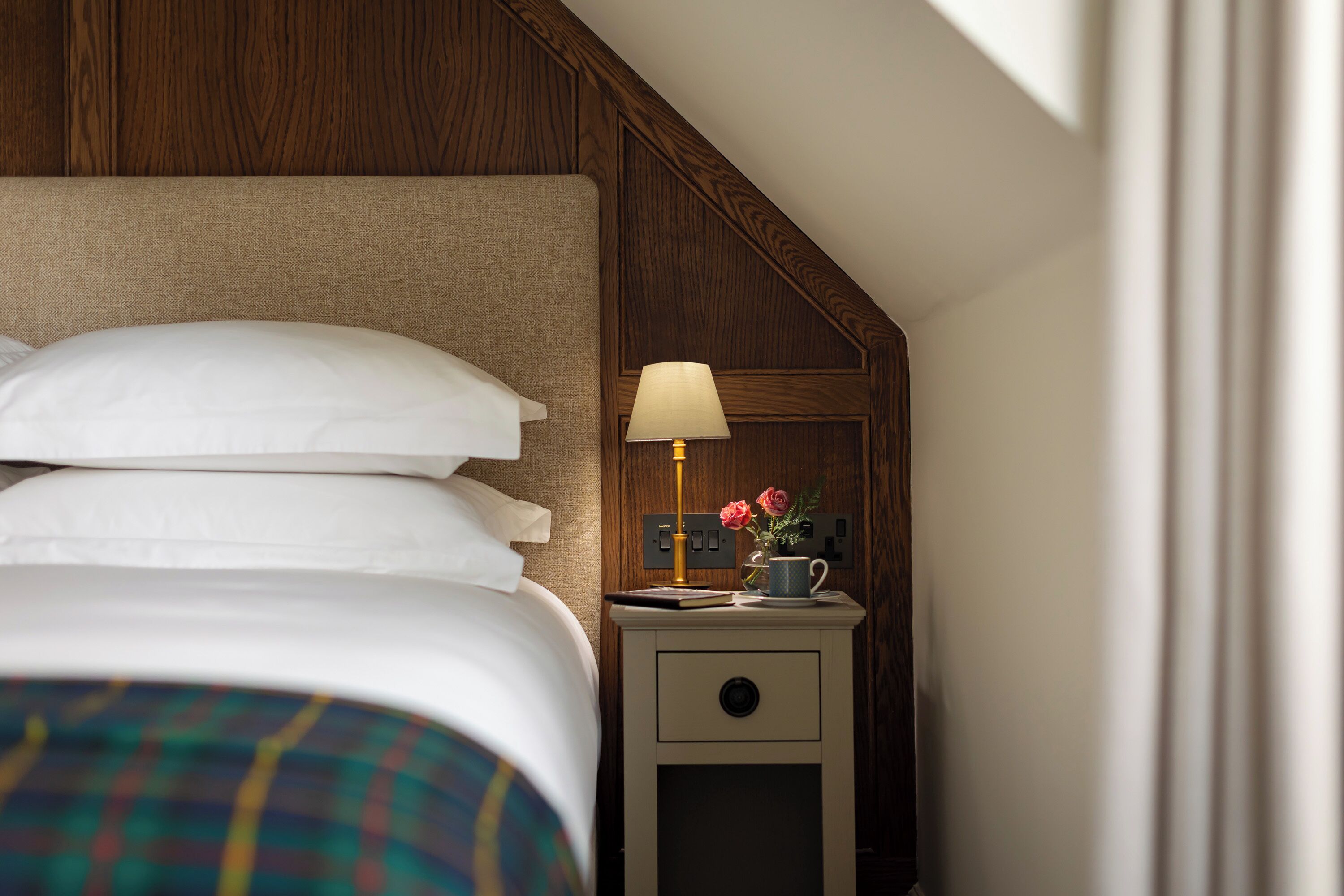 A neatly made bed with white linens and a plaid blanket, a bedside table with a lamp, a cup, and a vase with flowers, and a wooden panel wall.