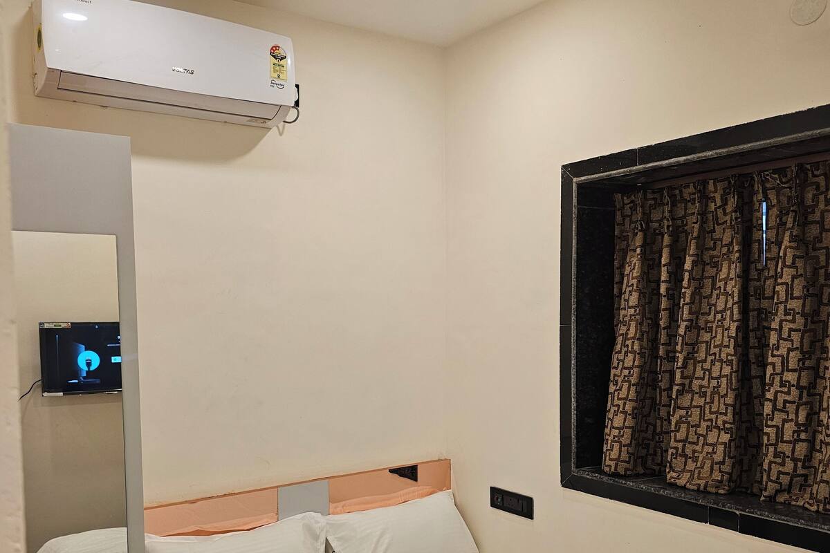 A hotel room with a bed, a window with patterned curtains, an air conditioning unit, a ceiling fan, and a small TV on a shelf.