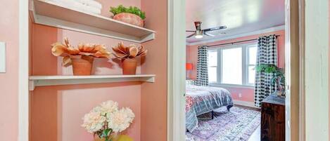 Un dormitorio con cama, alfombra, ventana con cortinas y estante con toallas y plantas.