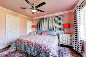 Un dormitorio con cama, ventilador de techo, cortinas estampadas y mesita de noche con lámpara.