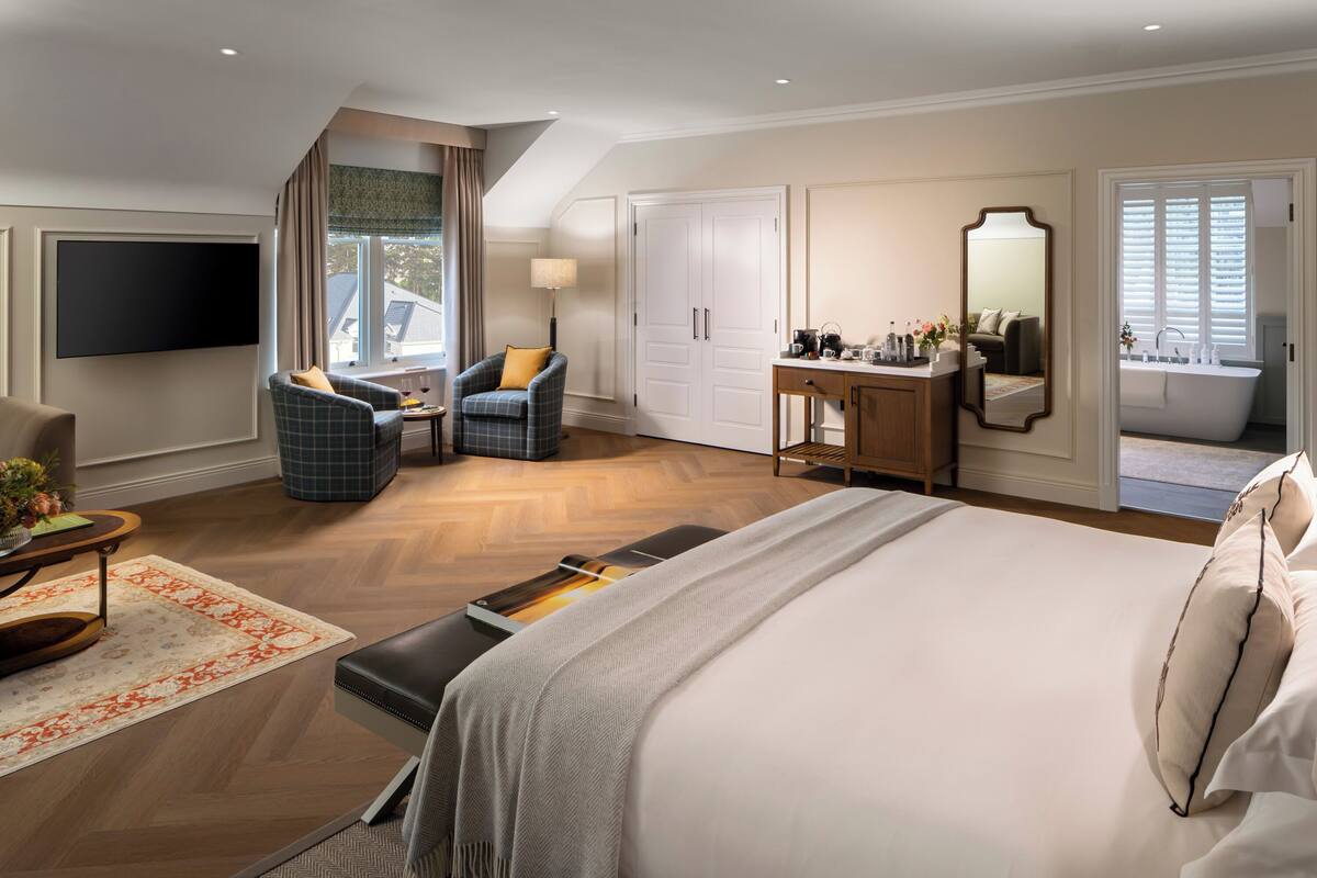 A modern hotel room with a large bed, a seating area with a chair and table, a desk with a mirror, a bathroom with a bathtub, and a TV mounted on the wall.