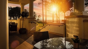 Un espace extérieur abrité, aménagé avec une table et des chaises, un pot de fleurs et offrant une vue sur le coucher de soleil derrière les arbres.