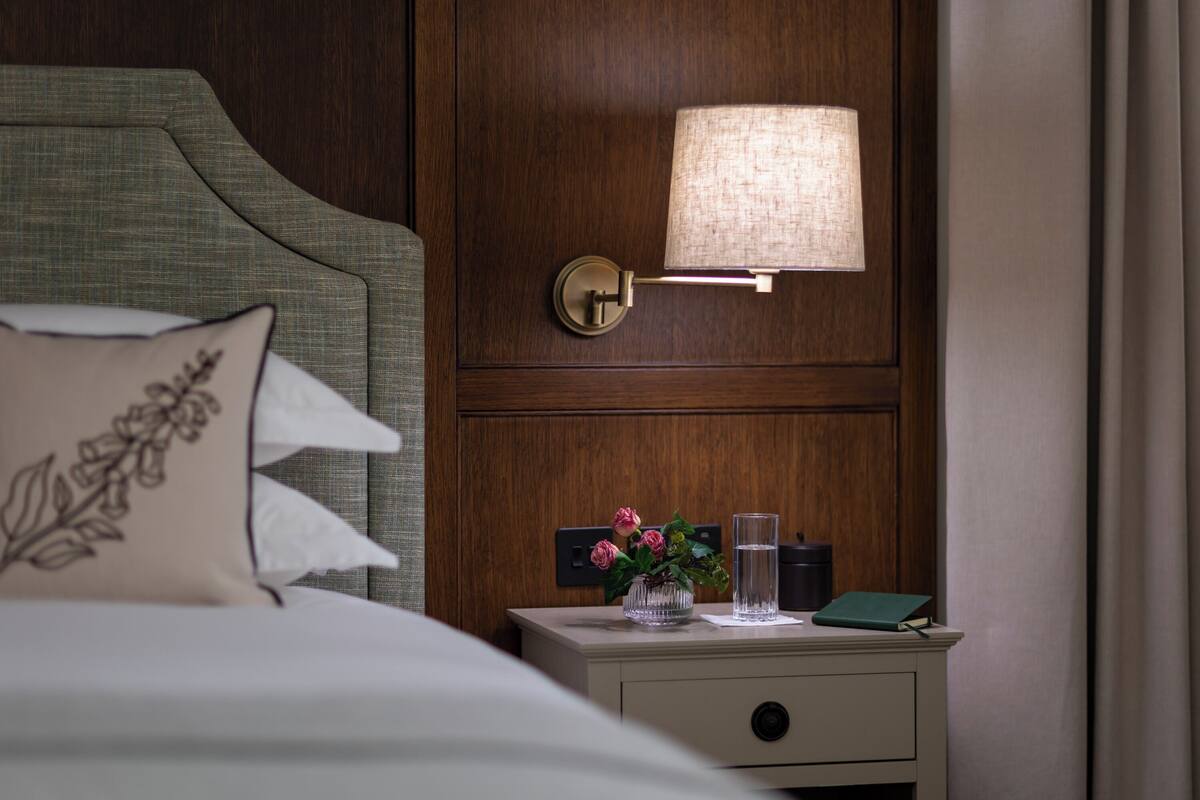 A neatly made bed with a pillow, a bedside table with a lamp, a vase with flowers, and a book.