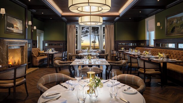 Une salle à manger élégante, avec plusieurs tables dressées pour le service, une cheminée et une vue sur l’extérieur.