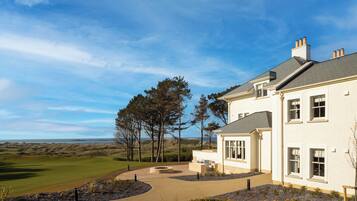 A large white house with a driveway leading to a landscaped area, surrounded by trees and a view of the sea.