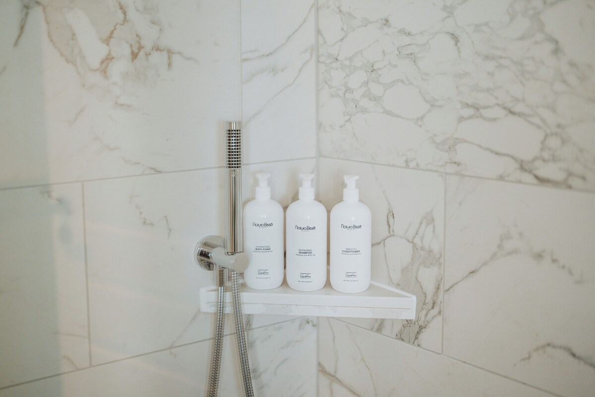 A bathroom with a marble wall, a shower, and three bottles of NaturaBisse products on a shelf.