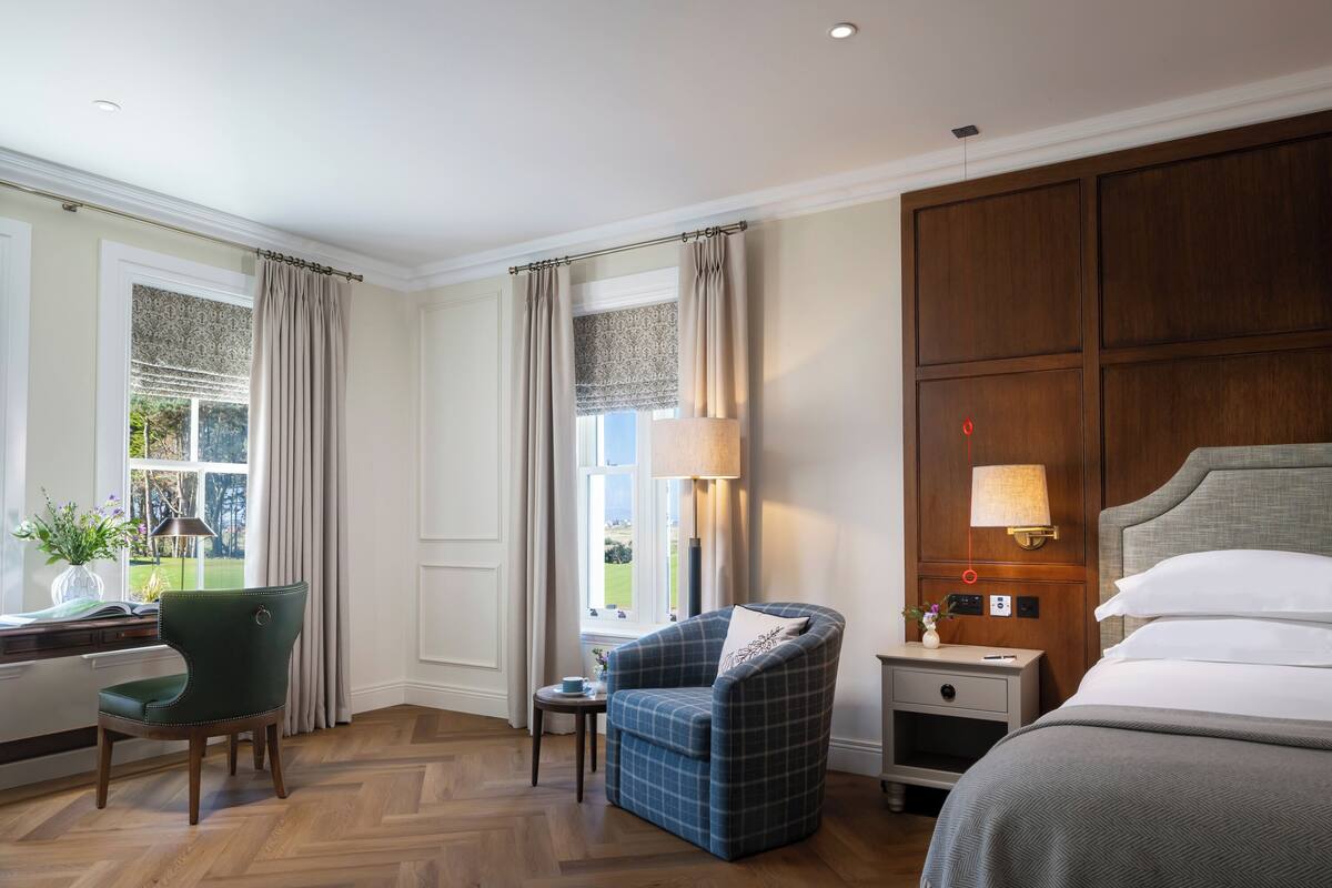 A hotel room with a bed, a desk, a chair, a nightstand, and a window with curtains.