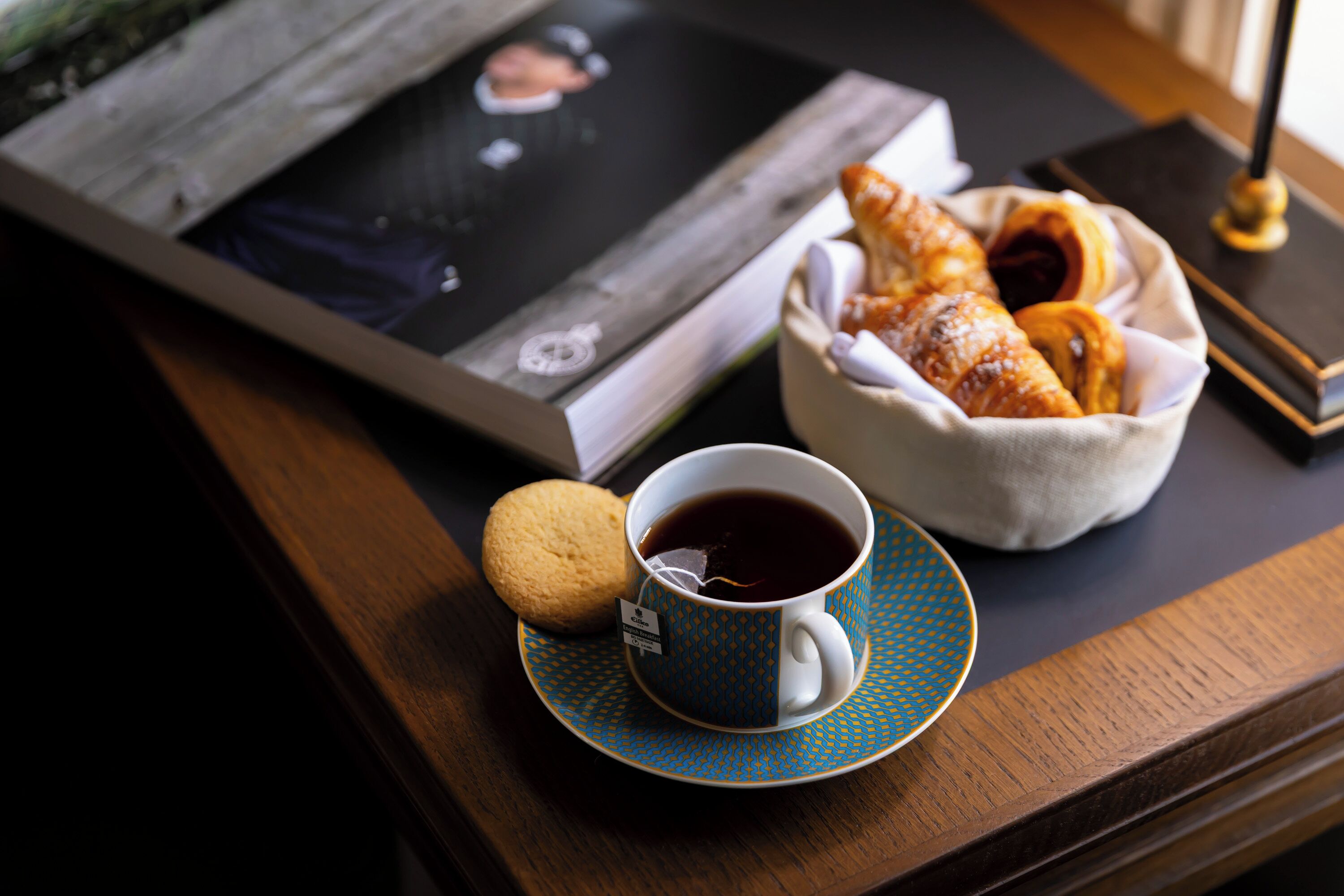 Une tasse de café accompagnée d’un biscuit sur un plateau, un panier de viennoiseries et un magazine sur une table en bois.