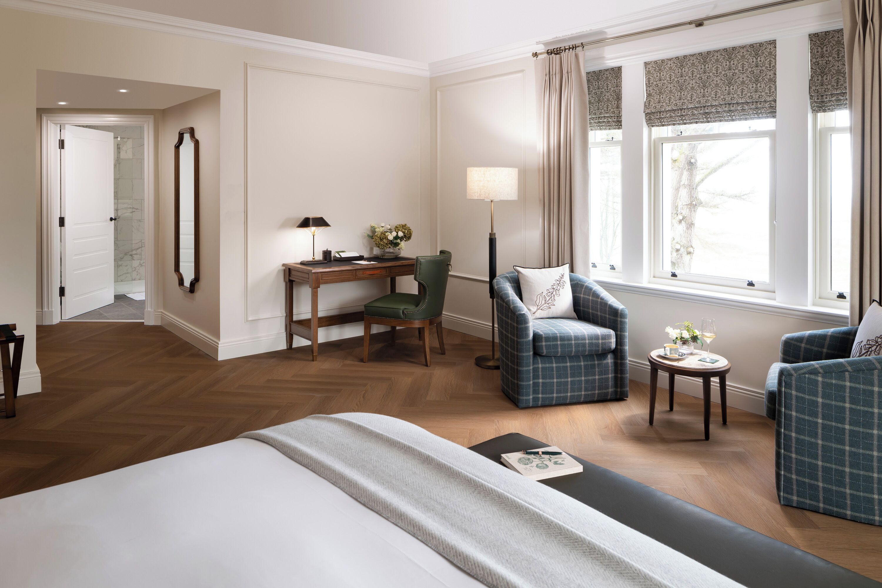 A hotel room with a large bed, a desk with a chair, two armchairs, a small table with a lamp, and a window with curtains.
