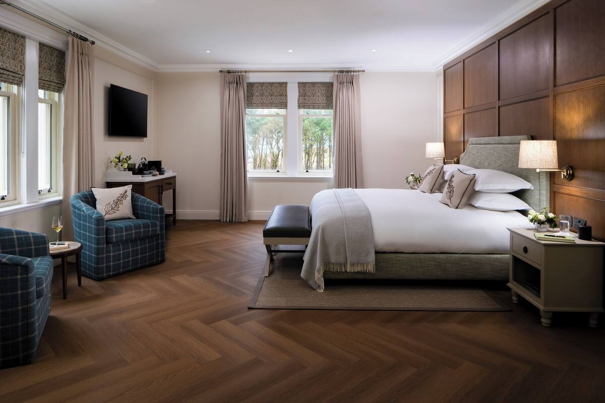 A bedroom with a large bed, a bedside table, a chair, a desk with a flat-screen TV, and a window with curtains.