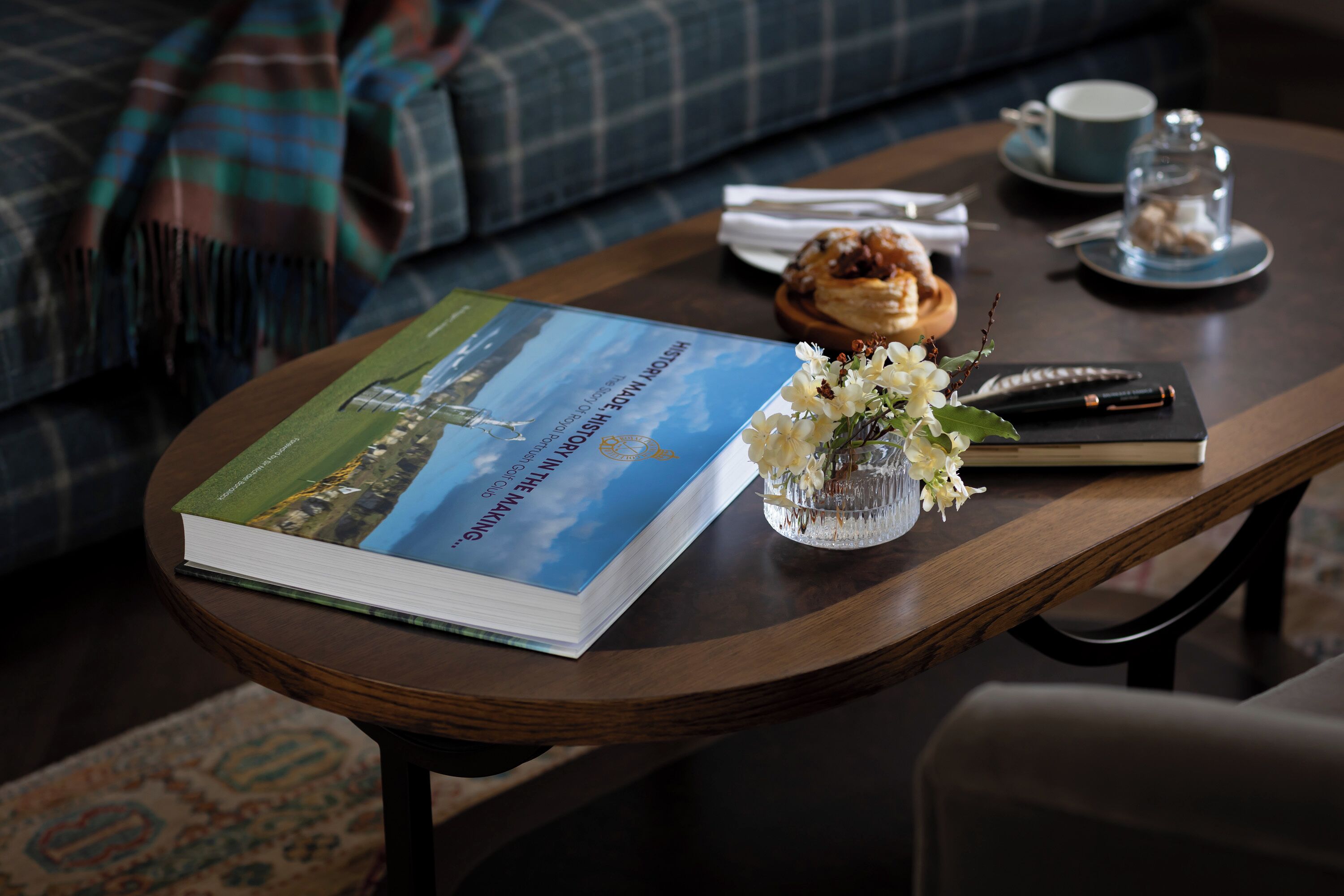 A book titled "The History of the Manhattan" on a wooden table, accompanied by a cup, saucer, and a small vase with flowers.