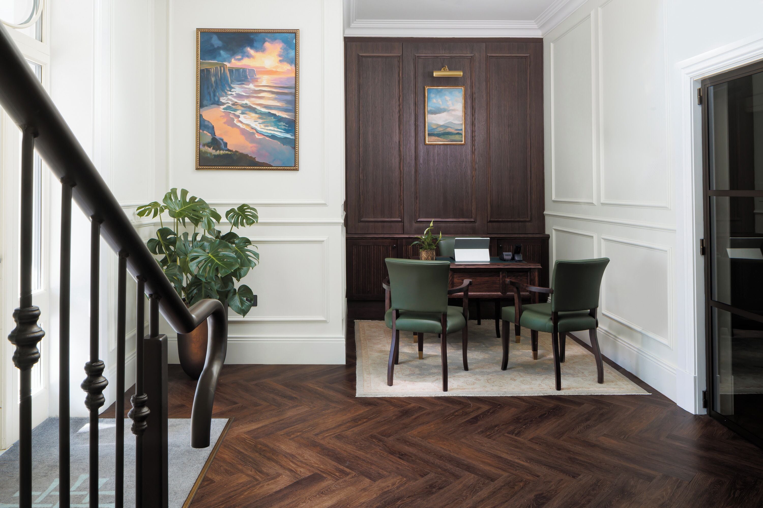 A hallway with a wooden floor, a staircase, a desk with chairs, and a painting on the wall.