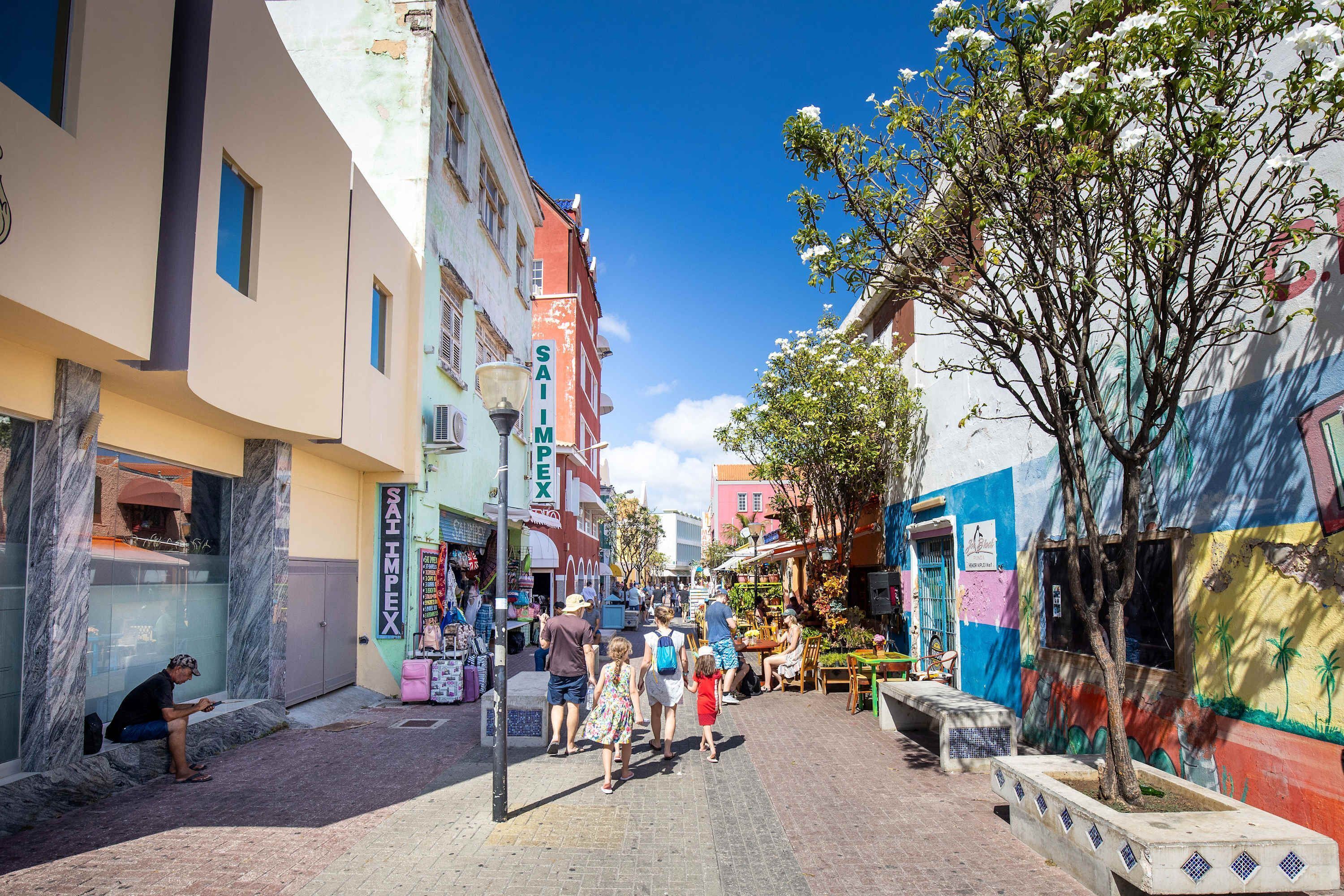 Une rue étroite bordée de bâtiments aux couleurs vives, un arbre aux fleurs blanches et quelques personnes qui se promènent.