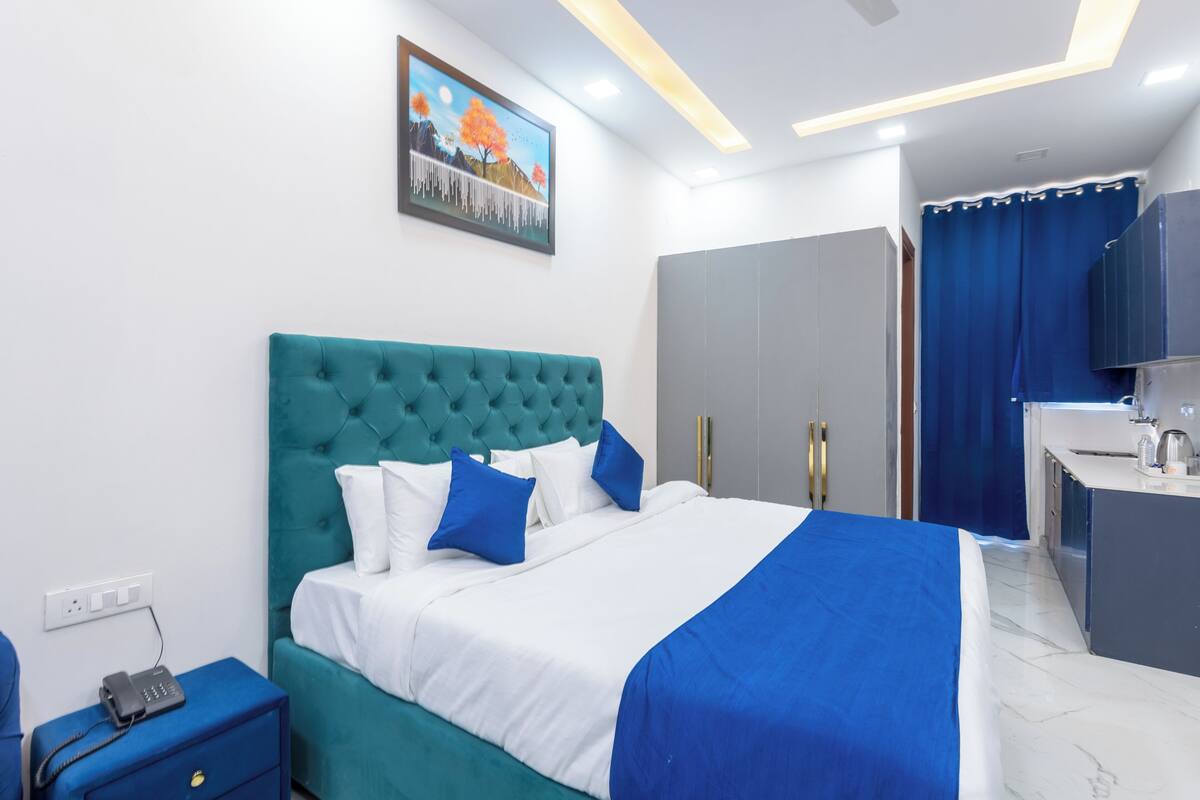 A modern hotel room with a teal upholstered headboard, a white and blue bedspread, a bedside table with a phone, and a painting of a landscape on the wall.