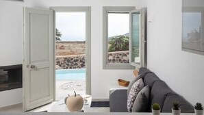 A room with a view of a pool and a stone wall outside. There is a sofa and a table with a vase and a bowl on it. - White Orchid Suites | 2 | Heated Shared Mini Pool (Vothonas)