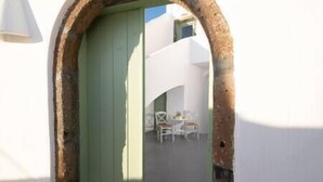 A narrow passageway with a green door, leading to a small outdoor seating area with white chairs and a table.