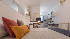 Un dormitorio con cama, almohadas, una silla, un escritorio, un televisor, un espejo y una kitchenette.