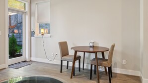 Un comedor con una mesa redonda y cuatro sillas, una ventana con vista a un muro de piedra y follaje, y un estante de pared con un reloj y una planta pequeña.