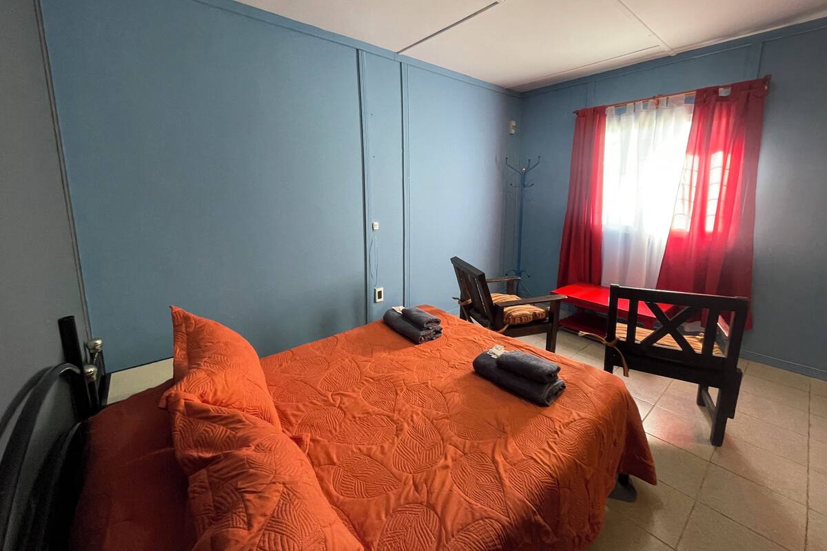 A room with two beds, a chair, a table, and a window with red curtains.