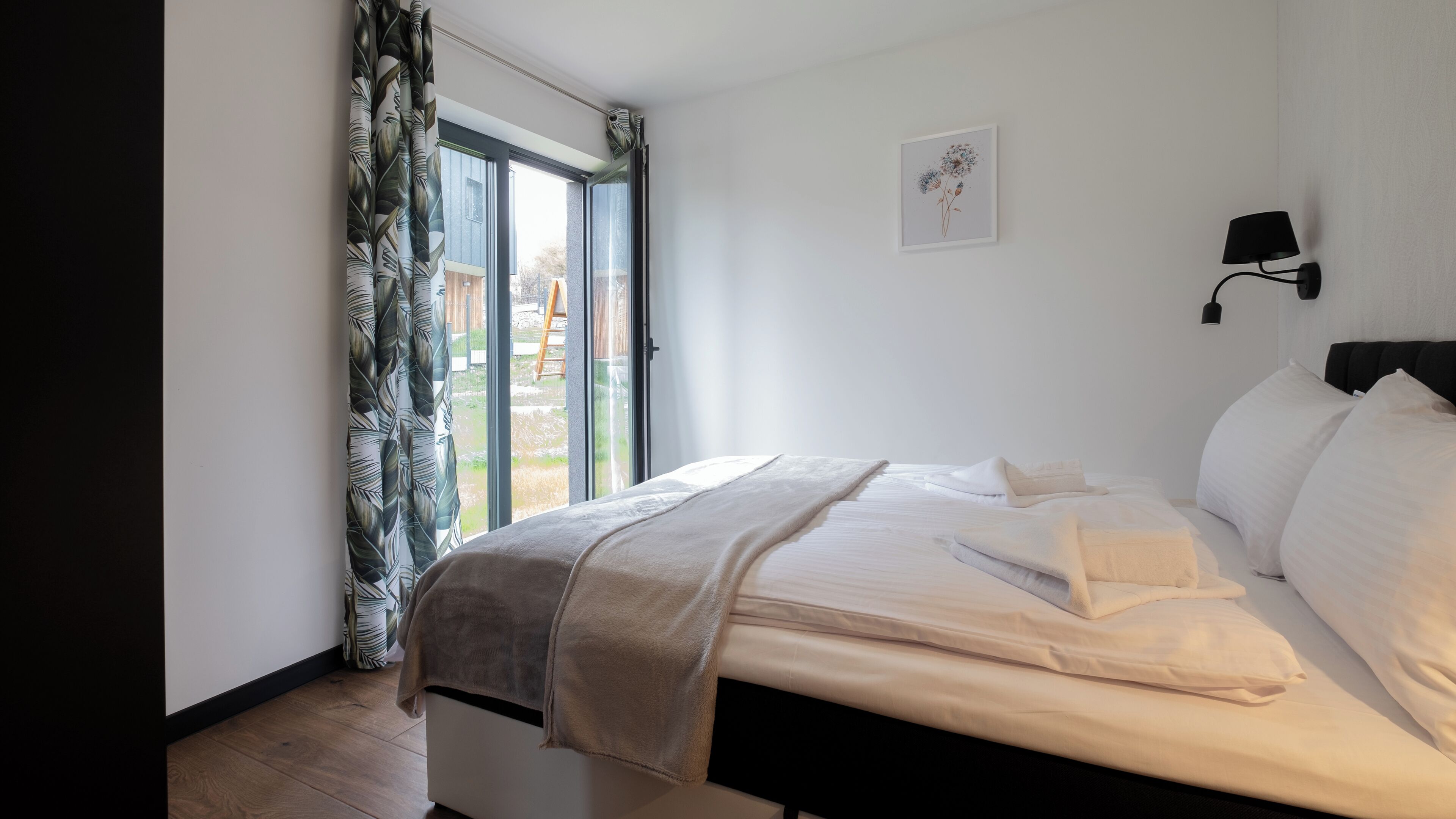 Ein Schlafzimmer mit Bett, einem Fenster mit Vorhängen, einer Wandlampe und einem gerahmten Bild.