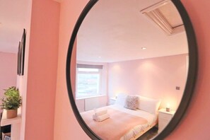 A bedroom with a large mirror, a bed with white linens, a bedside table, and a window.