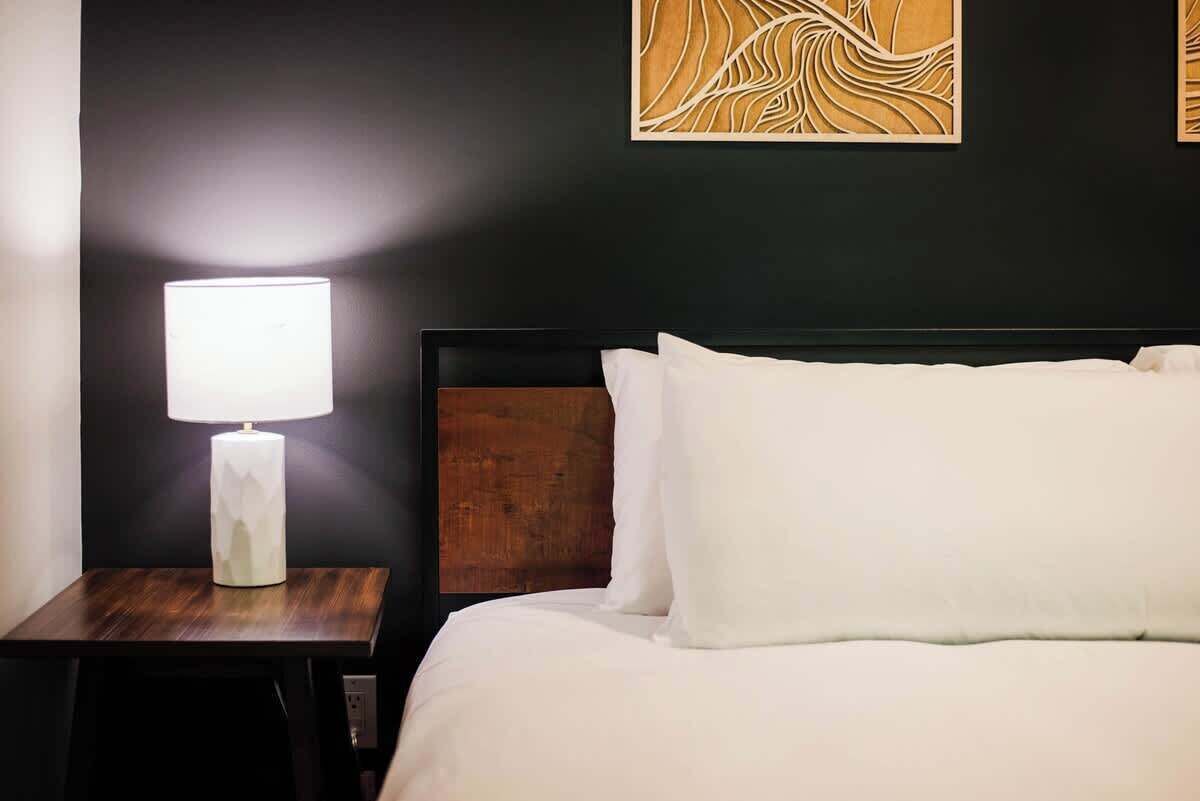 A neatly made bed with white linens and a wooden headboard. A small bedside table with a white lamp is visible.