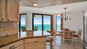 A kitchen with granite countertops, wooden cabinets, a dining area with a round table and chairs, and a view of the ocean through large glass doors.