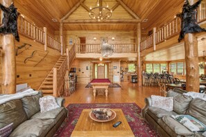 A spacious log cabin interior with a high ceiling, wooden floors, a billiards table, and a cozy seating area.