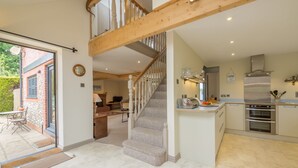 A spacious kitchen with a central island, a built-in oven, and a stainless steel range hood. A wooden staircase with a carpeted landing leads to an upper level. - Plumtrees (4), Thornham, Norfolk (Thornham)