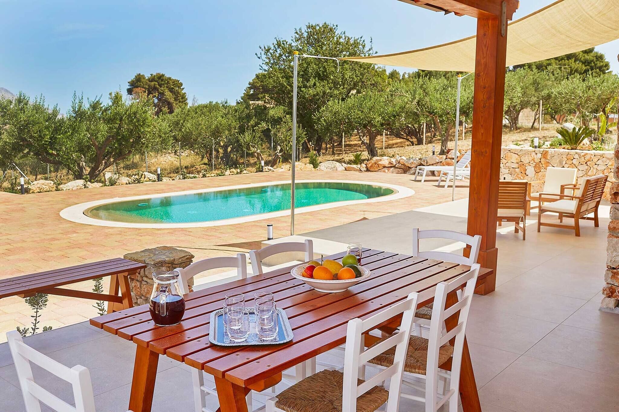 An outdoor pool area with a wooden table, chairs, and a fruit bowl.