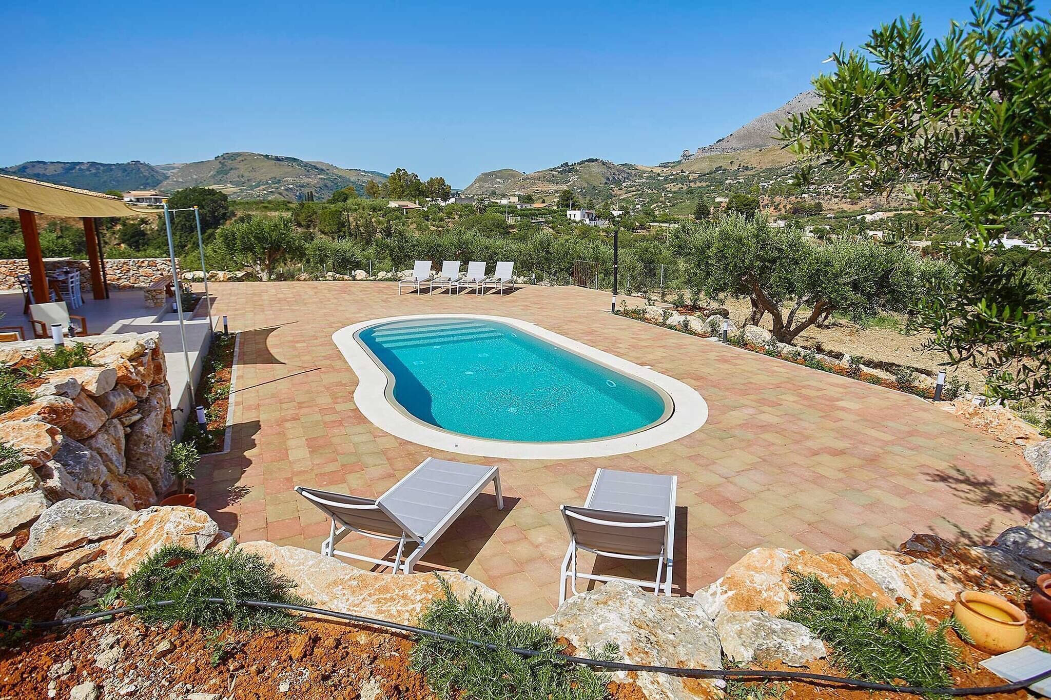 A swimming pool with lounge chairs, surrounded by a stone wall and vegetation, with a mountainous backdrop.