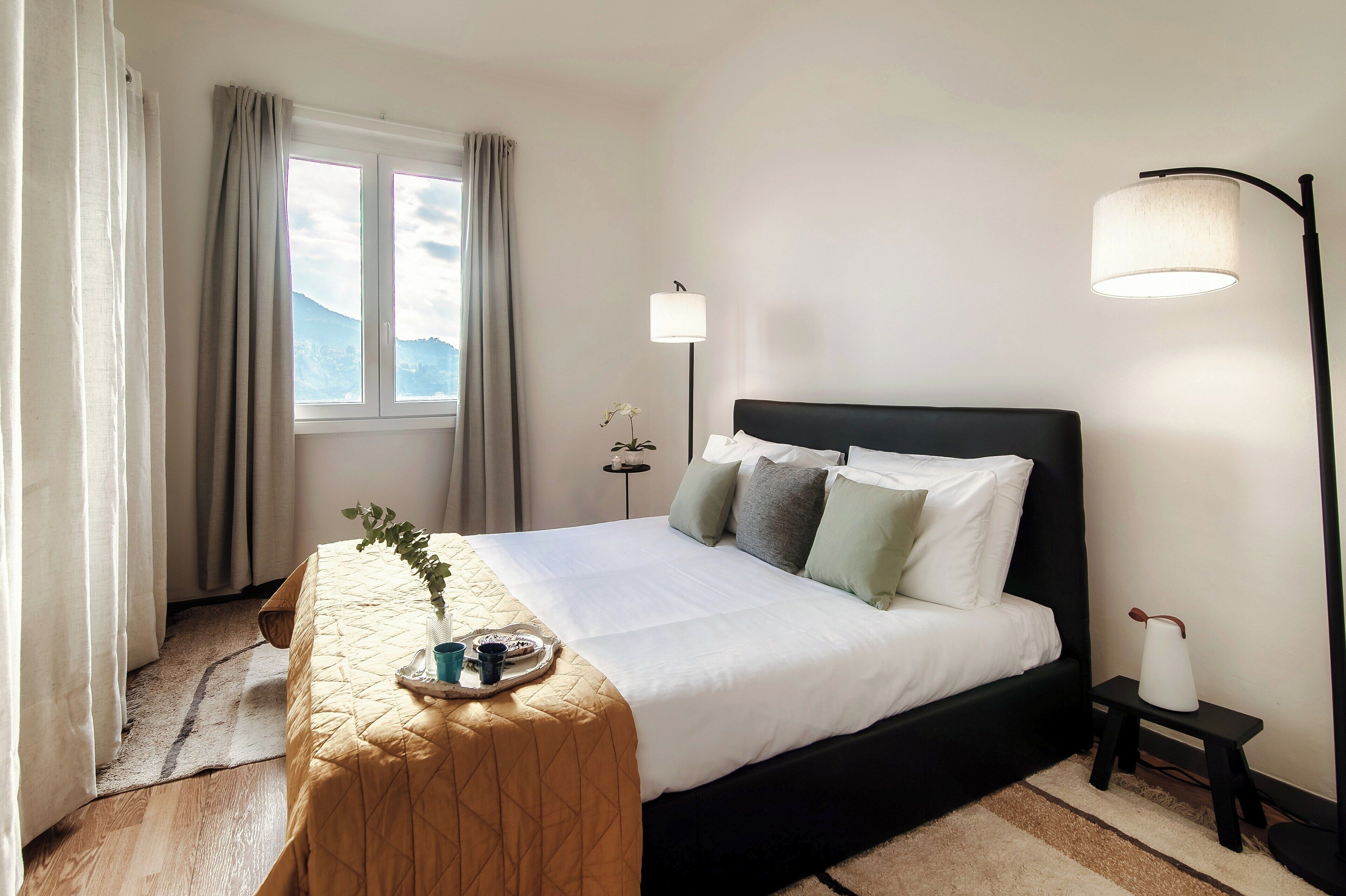 Un lit bien fait, avec une couette et des oreillers blancs, une table de chevet avec une lampe, et une vue sur l’extérieur par une fenêtre.