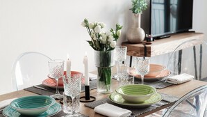 Ein Esstisch ist gedeckt mit klarem Glas, grünen und rosa Tellern und einer Vase mit weißen Blumen.
