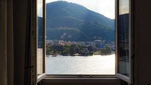 Blick durch ein offenes Fenster auf einen Fluss, Gebäude und eine Hügelkulisse im Hintergrund.