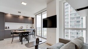 A modern kitchen with dark cabinets, a white tiled backsplash, and a dining area with a round table and chairs. A flat-screen TV is mounted on the wall above the dining area, and there is a large window providing a view of a high-rise building.