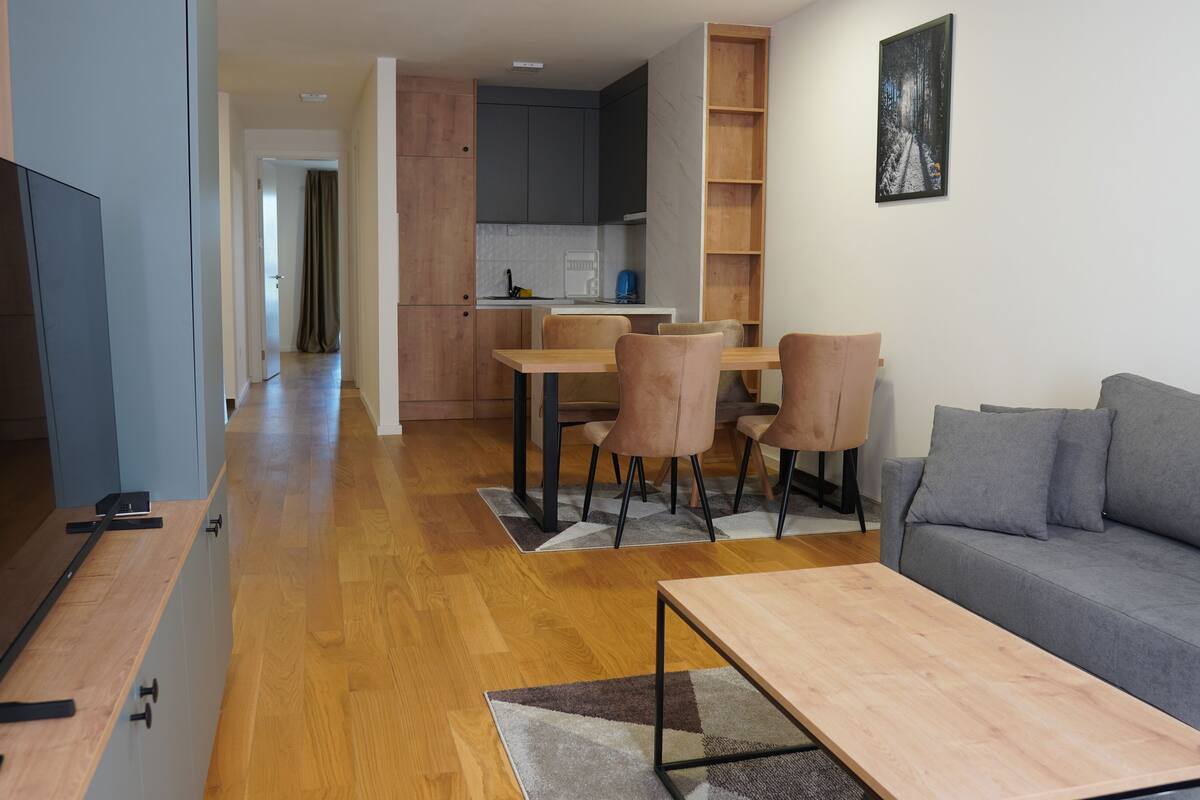 Una sala de estar moderna con un sofá gris, una mesa de centro de madera y un televisor sobre un mueble. Se observa un comedor con una mesa y sillas, y una cocina al fondo.