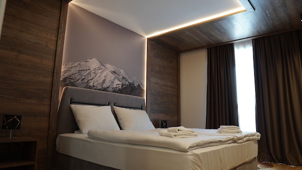 A modern hotel room with a large bed, wooden paneling, a mountain mural, and a large window with curtains.