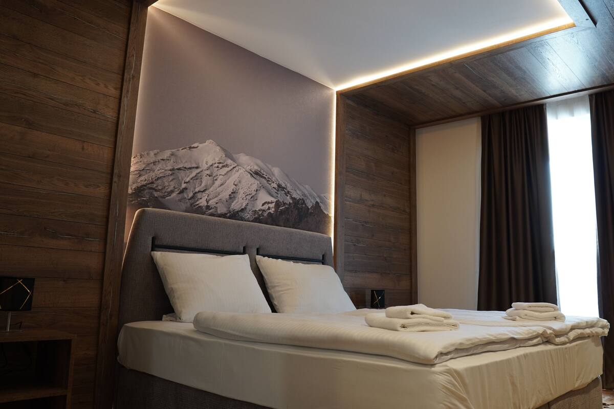 Una habitación de hotel moderna con una cama grande, paneles de madera, un mural de montañas y un amplio ventanal con cortinas.