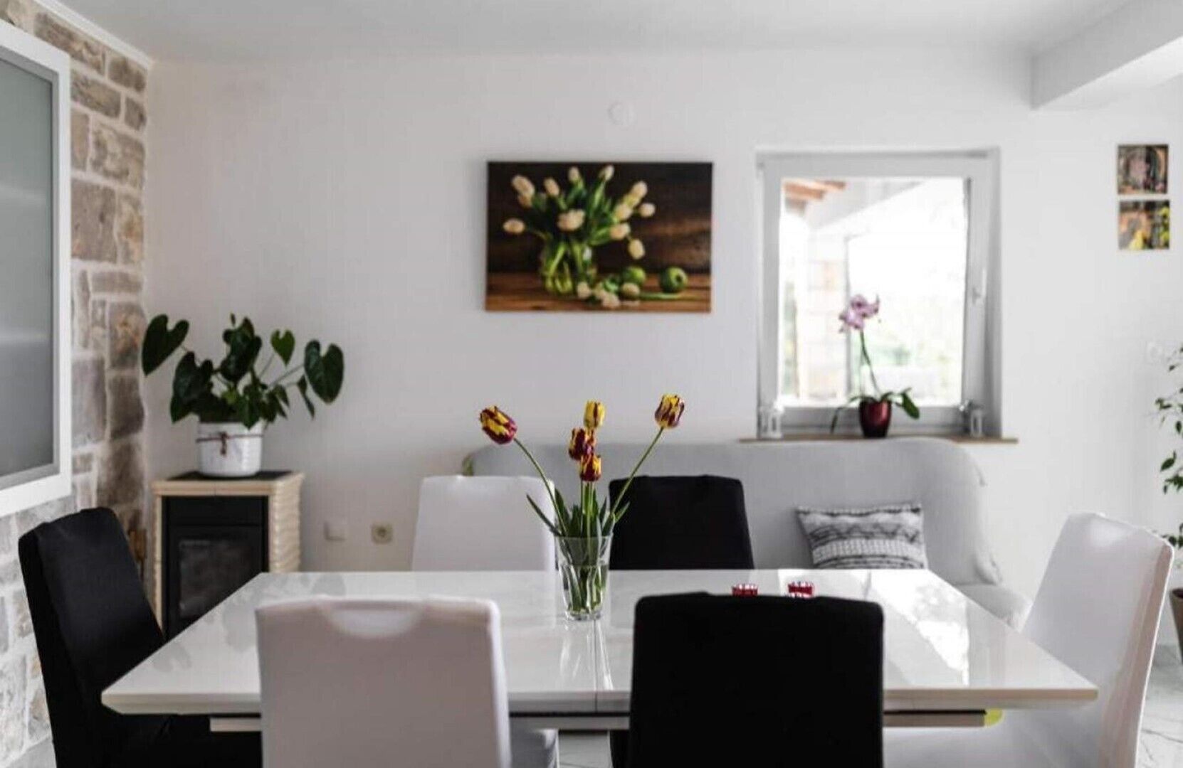 Un espace repas moderne avec une table blanche, des chaises noires et un vase de fleurs. On y trouve également une cheminée, une plante et une fenêtre donnant sur un beau paysage.
