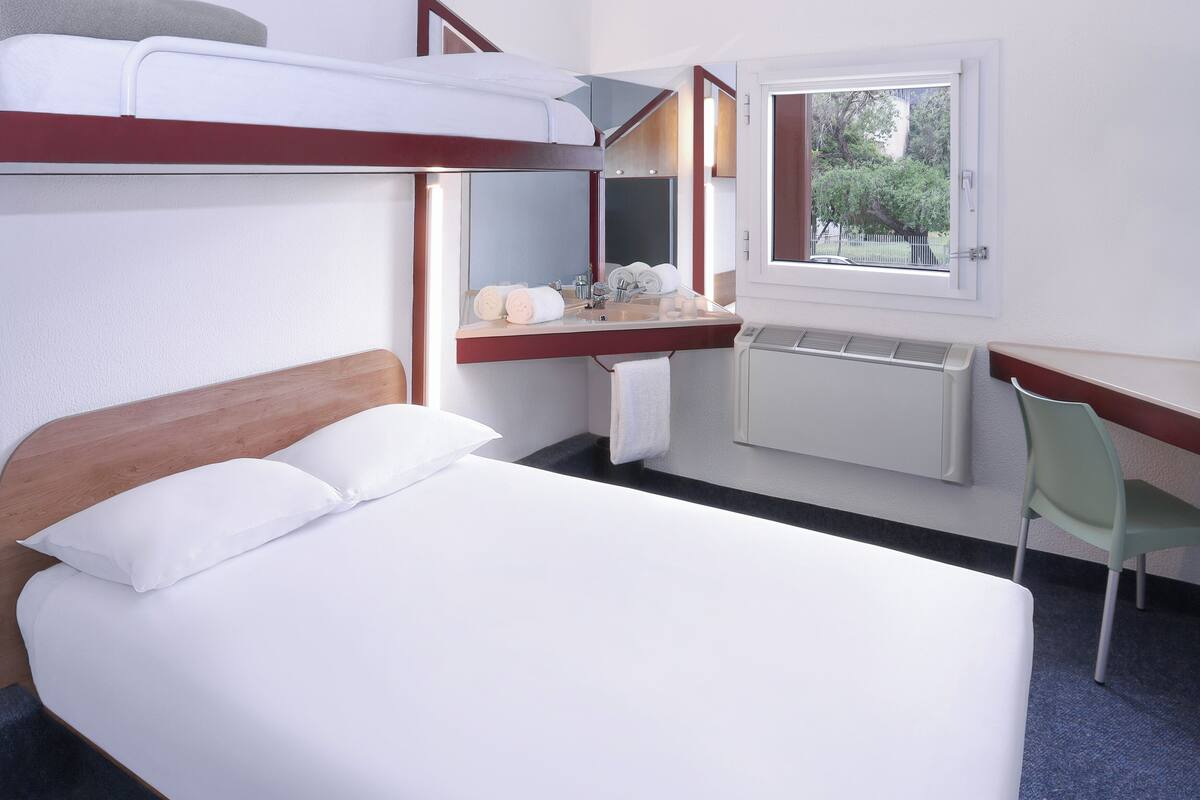 A hotel room with a bunk bed, a small desk with a chair, a window with a view of trees, and a radiator.