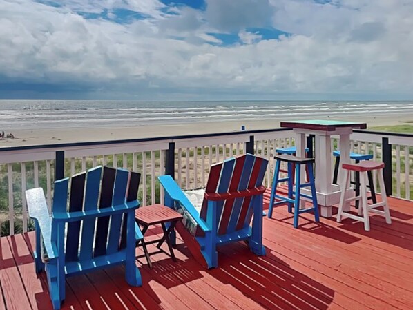 Terraza de madera con sillas Adirondack azules y rojas y una pequeña mesa, con vista a la playa.