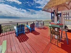 Una terraza de madera con sillas Adirondack azules y una mesa, con vista a una playa y un cielo despejado.