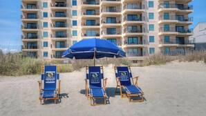 A beachfront hotel with multiple balconies, a sandy beach, and three blue lounge chairs under a blue umbrella. - 3BR/3BA Close to Main St-Oceanfront-Tidemaster 301 (North Myrtle Beach)
