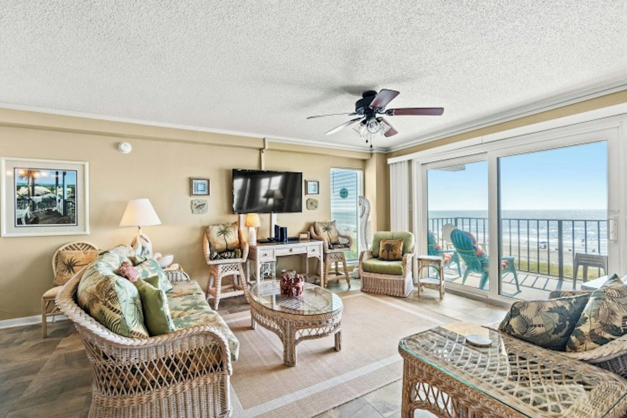 A spacious living room with wicker furniture, a ceiling fan, a flat-screen TV, and a large sliding glass door leading to a balcony with a view of the ocean.
