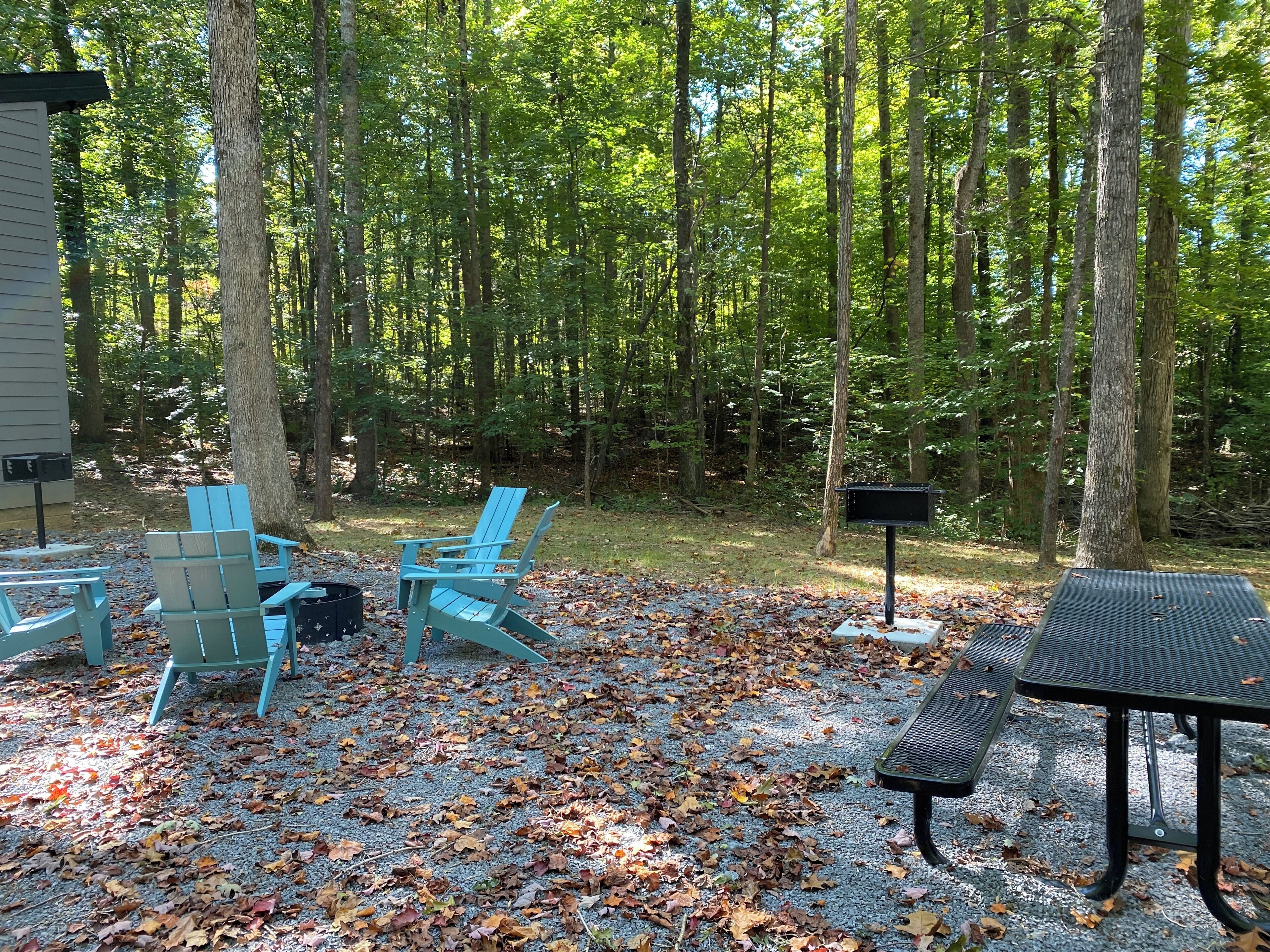 Een terras buiten met Adirondack-stoelen en een picknicktafel, omgeven door een bos.