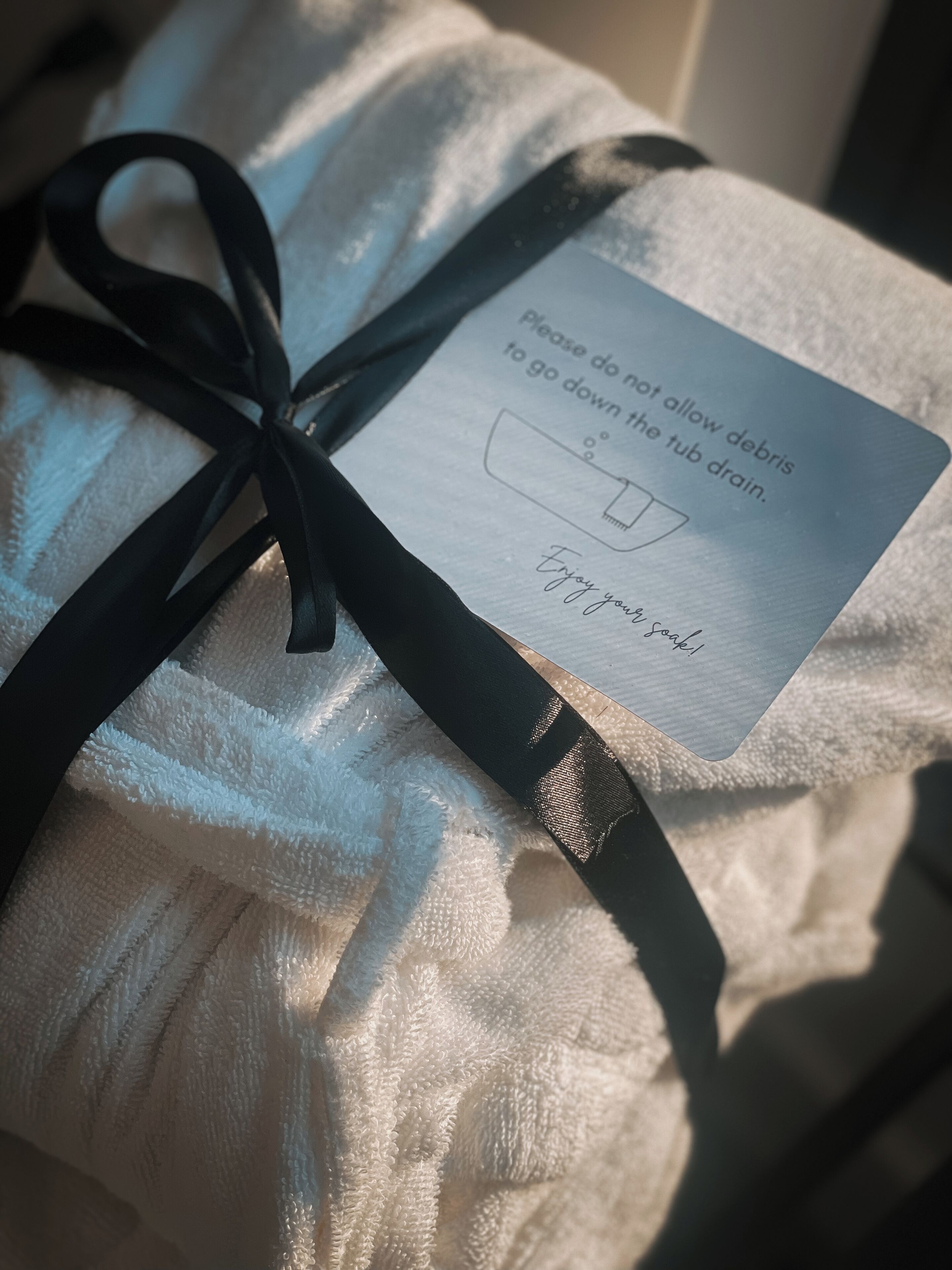 Une boîte cadeau ornée d’un ruban noir, contenant un mot de conseil : ne laissez pas de débris s’écouler dans le drain de la baignoire et profitez de votre bain.