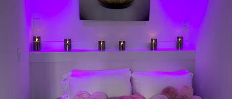 Un dormitorio con cama blanca, almohadas mullidas, una lámpara en forma de corazón montada en la pared y una obra de arte enmarcada con labios.