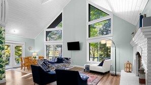 A spacious living room with a high ceiling, large windows, a fireplace, and a flat-screen TV. - Lakefront Spacious Cottage with Sandy Beach, Kayaks and Games! (Gaylord)