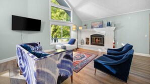 A living room with a fireplace, a flat-screen TV mounted on the wall, and a large window providing natural light. - Lakefront Spacious Cottage with Sandy Beach, Kayaks and Games! (Gaylord)